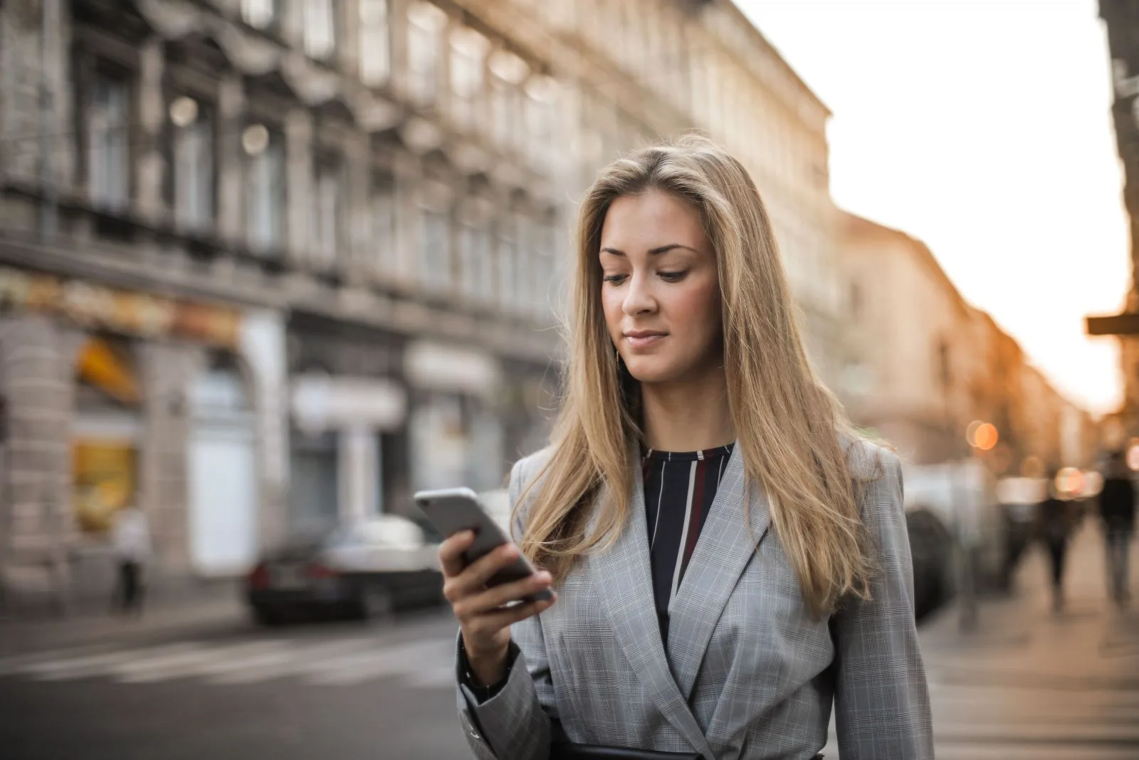 Mulher de blazer cinza olhando o celular enquanto caminha por uma rua urbana.