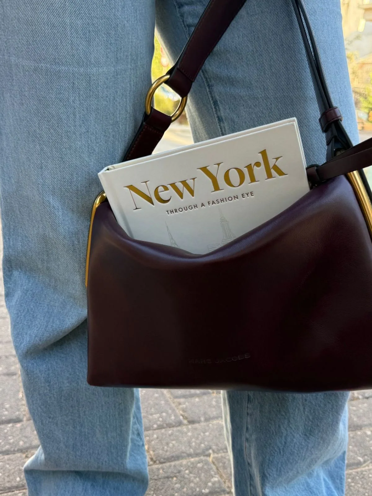 Pessoa usando jeans e bolsa marrom com um livro sobre Nova York dentro da bolsa