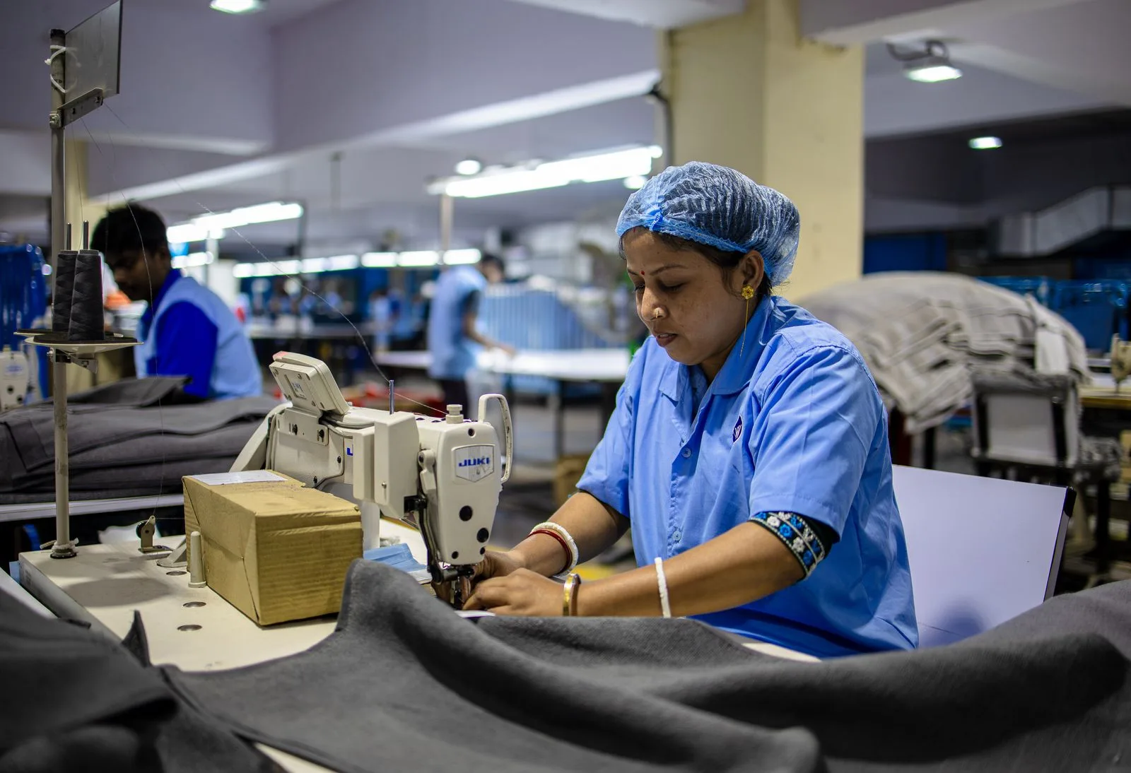 Trabalhadora operando uma máquina de costura em uma confecção de roupas, com tecidos e outros profissionais ao fundo.