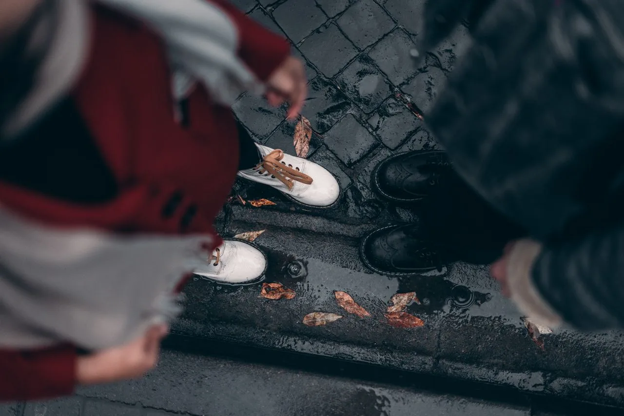 Pessoas usando calçados fechados em uma calçada molhada, em cena urbana com clima chuvoso.